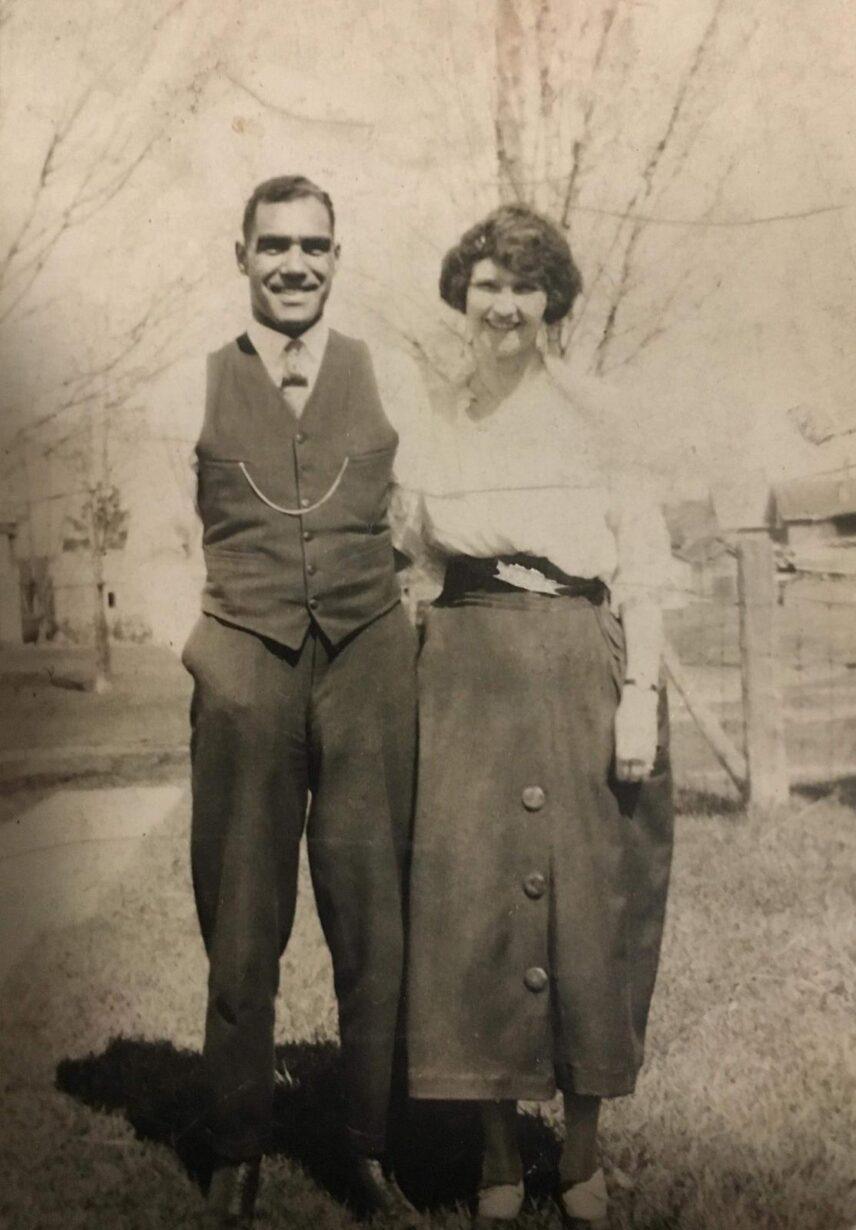 A black and white image of a man and woman. They are both standing and smiling. The man, who is on the left is wearing a suit vest with a pocket watch chain across it. The woman (on the right) is wearing a shirt and skirt with a black band around the center. She has her arm around the man.