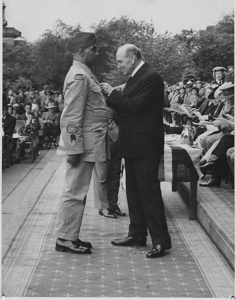 A black and white image of a two men surrounded by a seated crowd. The men are facing each other. The man on the left is wearing an army uniform. The man on the right is wearing a suit and is pinning a medal on the man on the left.