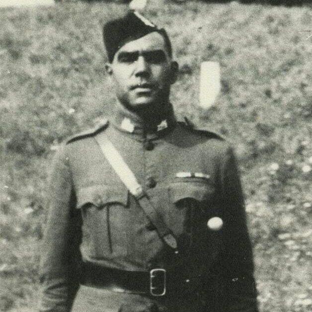 A black and white image of  a man standing with his arms by his side. He is wearing a regimental uniform that includes tartan pants.