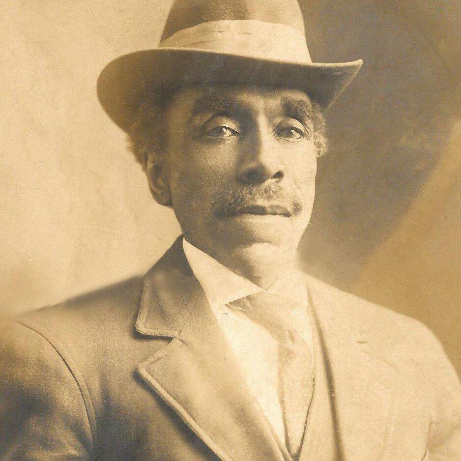 A sepia portrait of a man in a suit. He has a moustache and is wearing a hat.