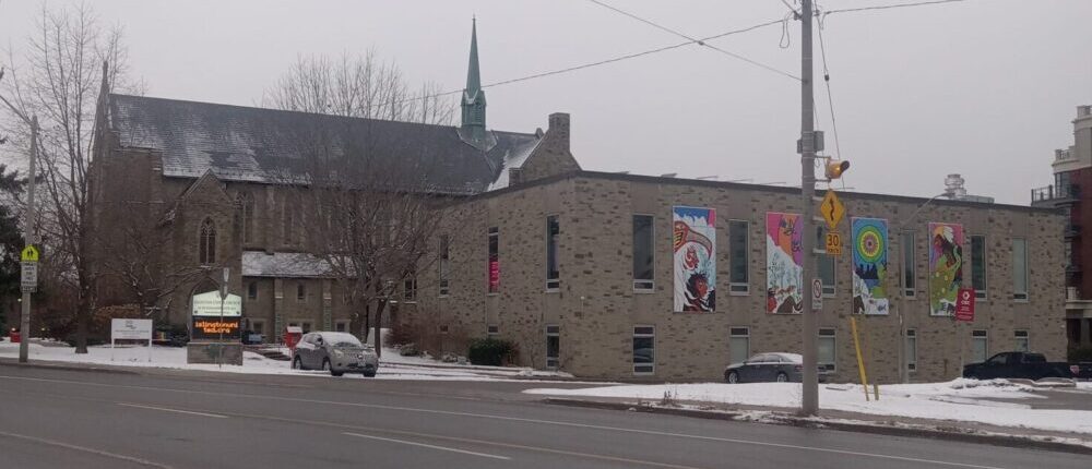 Une grande église en briques de couleur beige. L'église possède un petit clocher. A côté de l'église principale se trouve une aile supplémentaire construite avec des matériaux similaires.