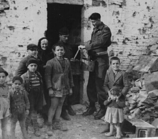 Image en noir et blanc d'un groupe d'enfants regardant l'appareil photo. Ils se tiennent devant un bâtiment en ruine. Derrière eux se trouvent une vieille femme et deux soldats. L'un des soldats tient deux conteneurs métalliques.