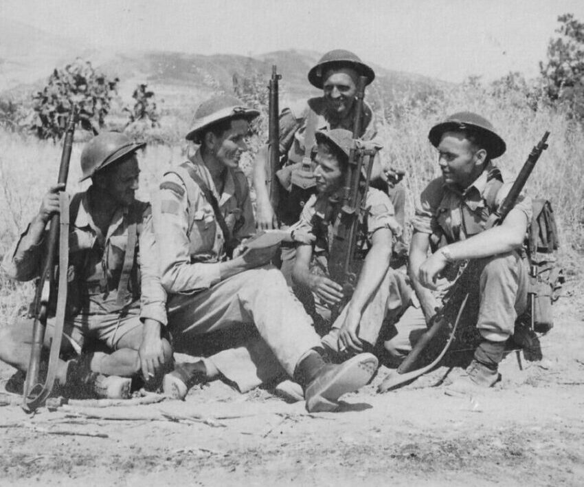 Image en noir et blanc d'un groupe de cinq hommes. Ils portent tous des uniformes militaires et sont assis par terre. Quatre d'entre eux regardent l'homme du milieu. Ce dernier tient un livre ouvert.