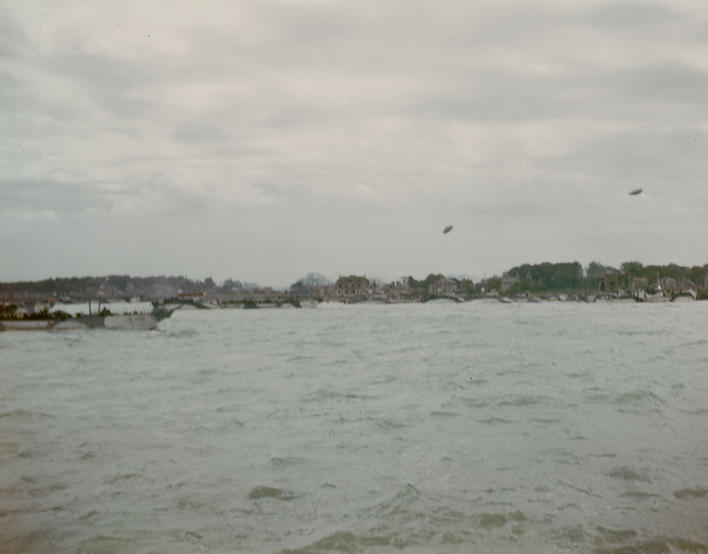 A view of a beach from the water. There is a military vessel in the water. The image is somewhat blurry. In the distance there are houses.