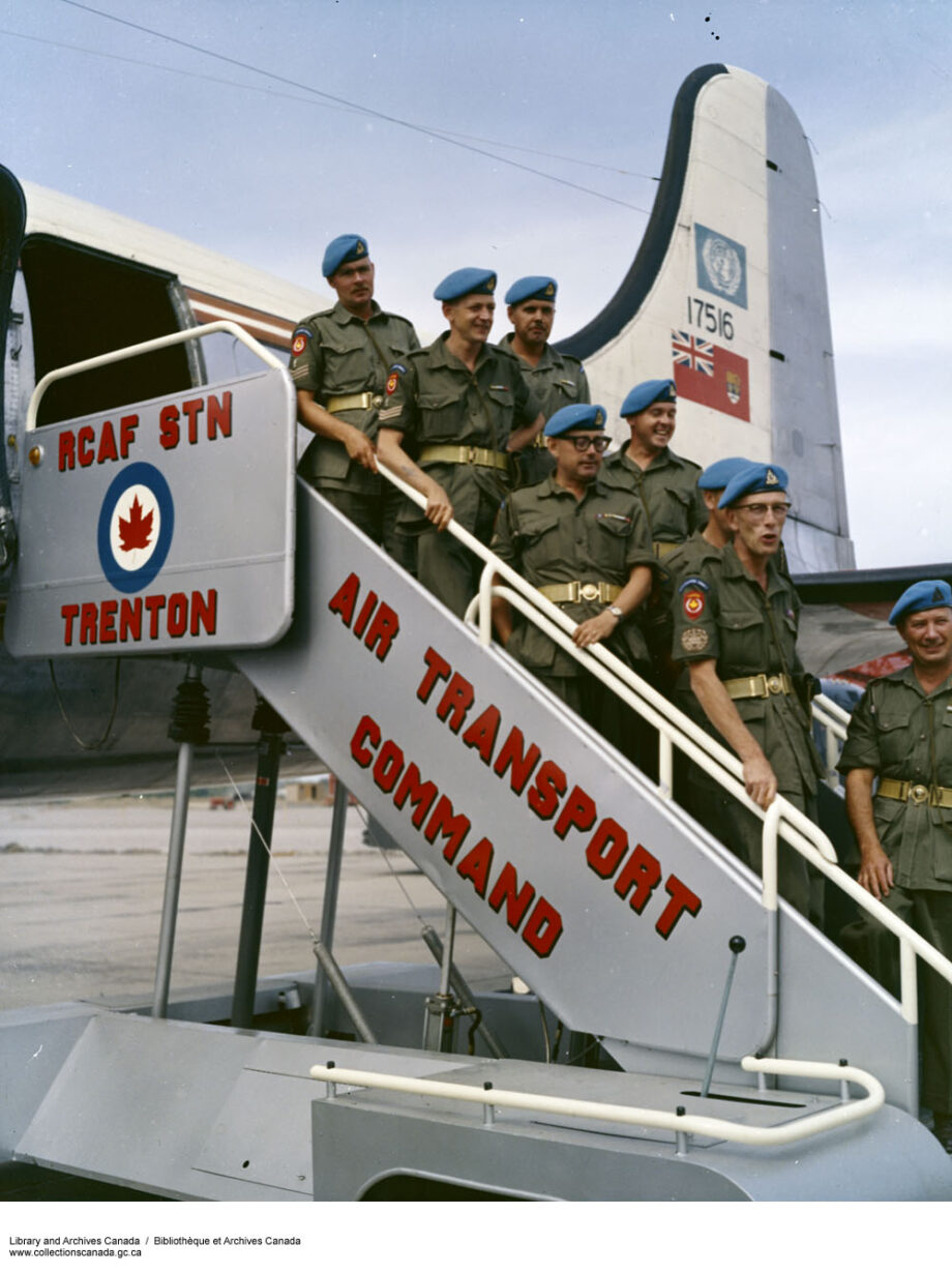 Un groupe d'hommes sur un escalier menant à un avion militaire. Ils portent des uniformes militaires et des bérets bleus. Sur l'empennage de l'avion figurent le logo des Nations unies et le drapeau rouge du Canada.