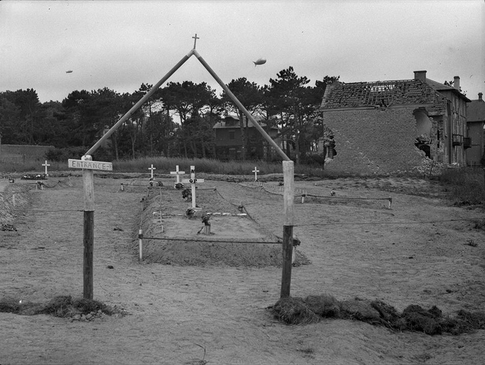 Image en noir et blanc d'un petit cimetière. Il y a des croix sur certaines tombes. À droite, un bâtiment partiellement détruit.