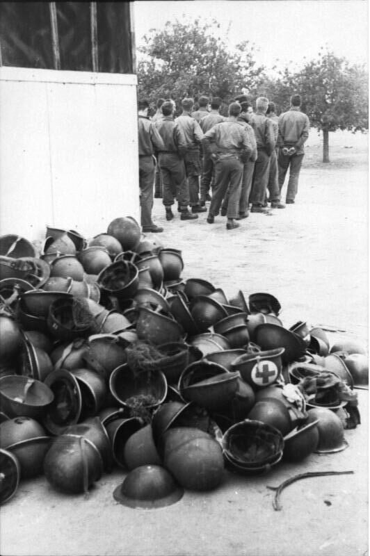 Une pile de casques à l'extérieur d'un bâtiment. À l'arrière-plan, un groupe de soldats fait face à la caméra.