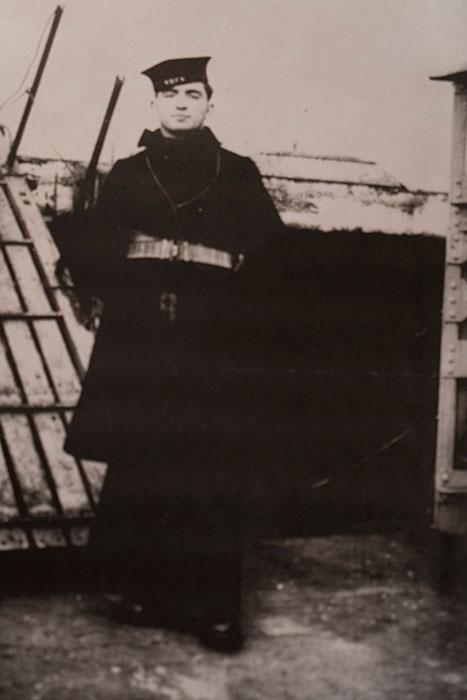 Un homme en uniforme de la marine. Il se tient debout, les mains derrière le dos, sur le pont d'un navire.