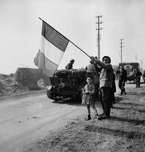 Un homme tenant un drapeau français au bord d'une route. À ses côtés, une jeune fille. Derrière lui, des soldats et des véhicules militaires circulent sur la route.