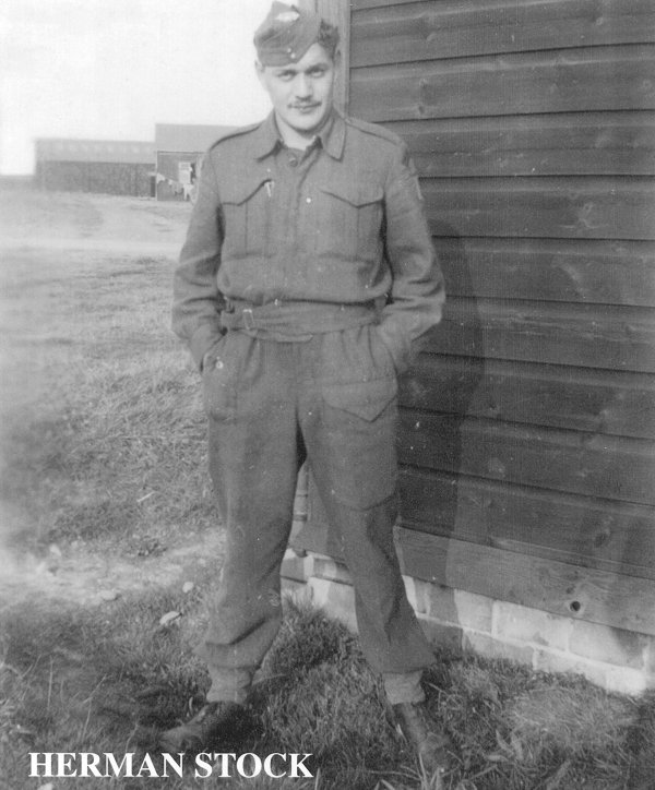 Un homme debout à côté d'un bâtiment militaire. Il porte un uniforme et a les mains dans les poches de son pantalon. Il porte une moustache.