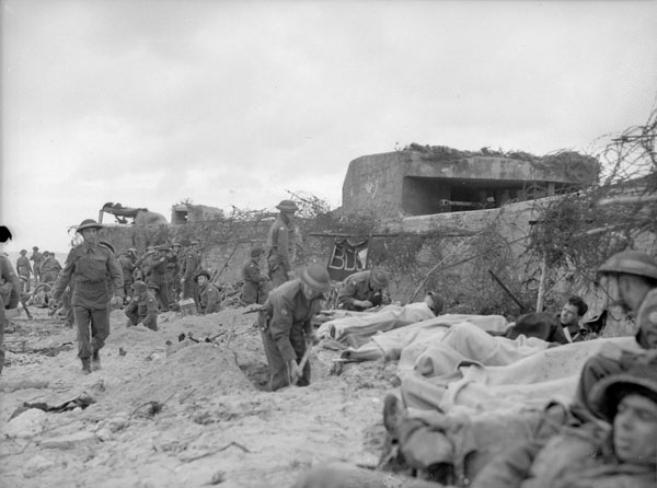 Un groupe de soldats sur une plage. Ils sont près d'un mur. Certains marchent, d'autres creusent. Sur le côté droit de l'image, un groupe de soldats blessés, vêtus de couvertures, sont soignés.