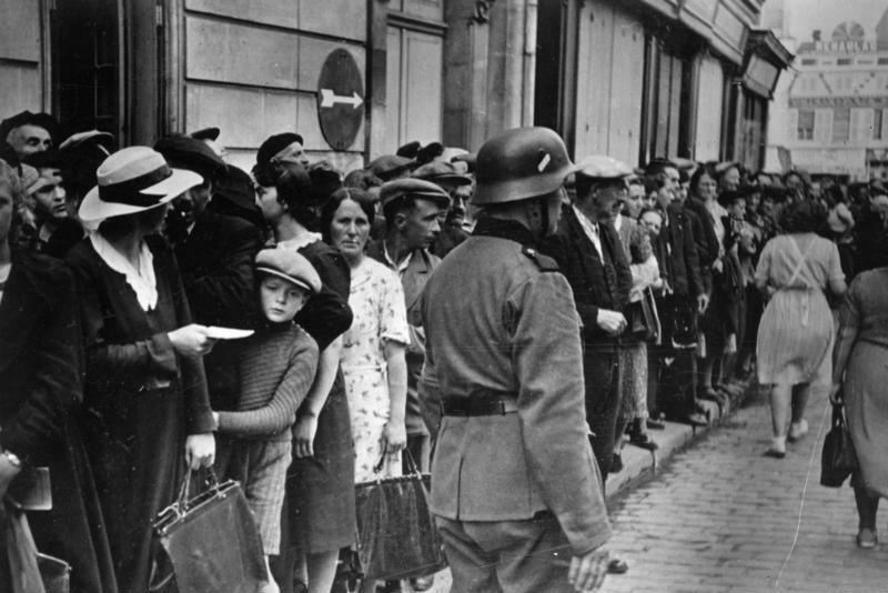 A group of people lining a street. There is a soldier beside them.