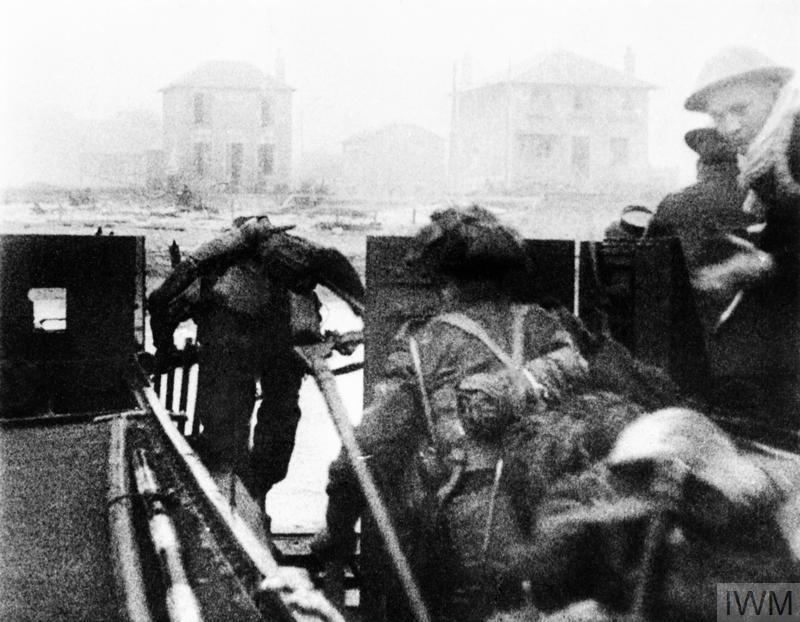 Image en noir et blanc de soldats quittant une péniche de débarquement. Ils se dirigent vers une plage. À l'arrière-plan se trouvent de grandes maisons.