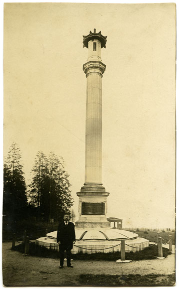 Un homme se tient devant un monument. Le monument est composé d'une haute colonne avec une base en pierre. Des inscriptions figurent sur la base du monument.