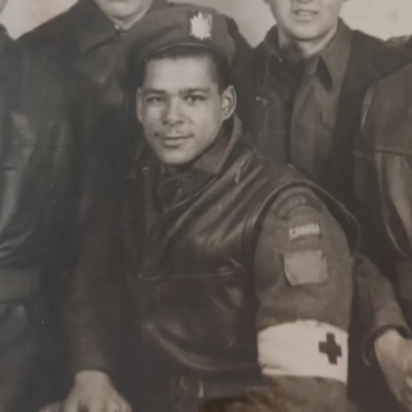 Image en noir et blanc d'un homme en uniforme militaire.Il est assis. Il porte au bras gauche un bandeau avec une croix. Derrière et autour de lui se trouvent d'autres hommes en uniforme.