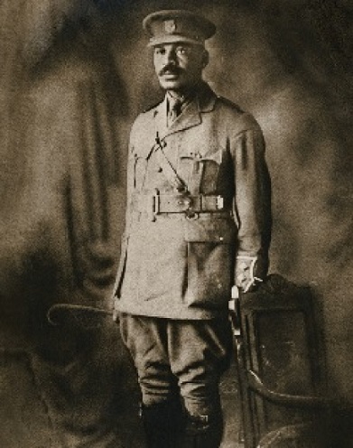 Image sépia d'un homme debout à côté d'une chaise, portant un uniforme militaire et une moustache.