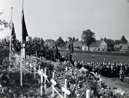 A man standing in a graveyard. In the graveyard are many flowers. In the background is a large group of people.
