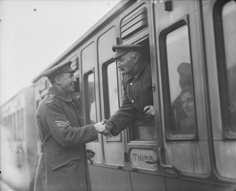 A man standing beside a train. He is shaking hands with another man inside the train through the window. They are both wearing military uniforms. 