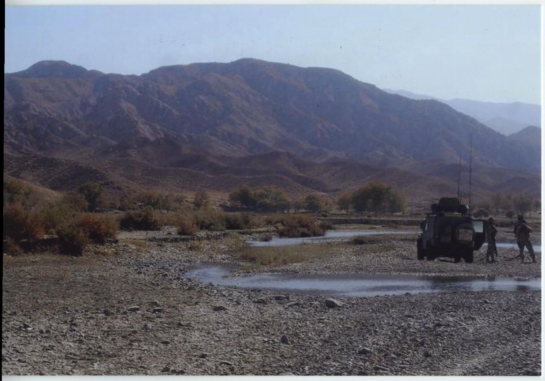 A hilly landscape in Afghanistan. In the foreground is a armoured vehicle and two soldiers.