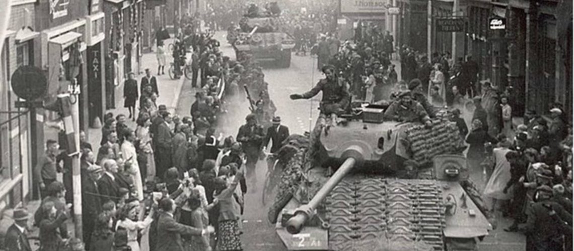 Un défilé de chars d'assaut dans une rue bondée de spectateurs. Sur les chars, des soldats saluent la foule.