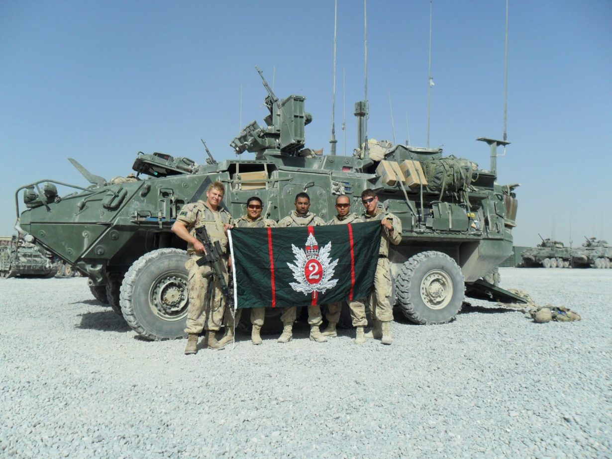 Un groupe de soldats devant un véhicule blindé léger. Ils tiennent un drapeau du Queen's Own Rifles of Canada.