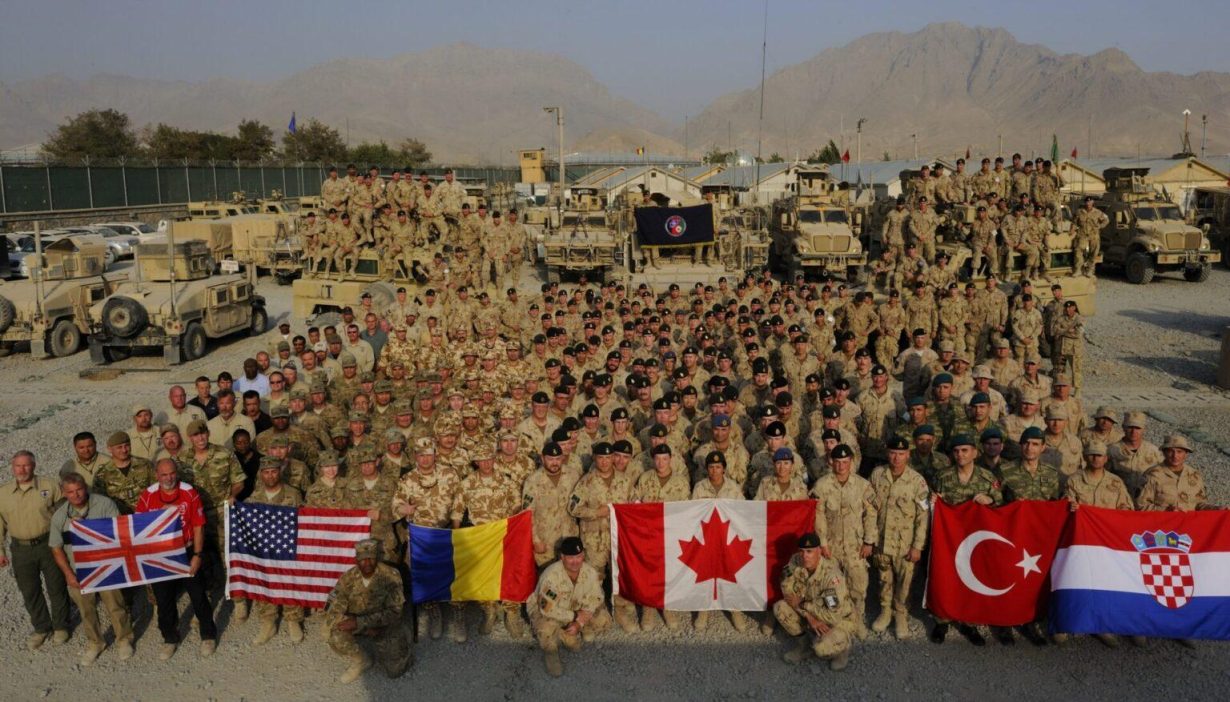 Un groupe de soldats en Afghanistan. Ils portent différents uniformes et brandissent les drapeaux du Royaume-Uni, des États-Unis d'Amérique, de la Roumanie, du Canada, de la Turquie et de la Croatie.