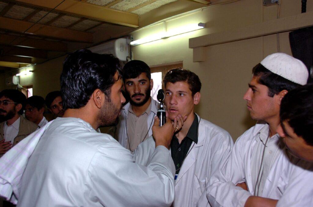 Un homme examine l'œil d'un autre homme à l'aide d'un instrument médical. D'autres hommes observent la scène.
