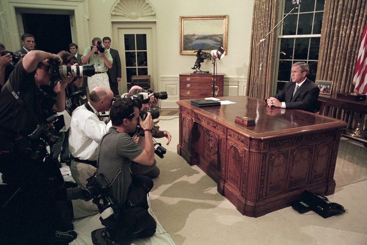 Le président George W. Bush assis à son bureau dans le bureau ovale. Plusieurs photographes le prennent en photo.