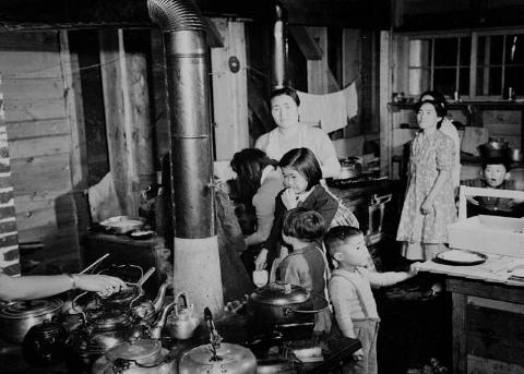 Un groupe de femmes et d'enfants dans une pièce à vivre construite principalement en bois. On y trouve un poêle à bois avec des casseroles et une table. Derrière eux, du linge est en train de sécher.