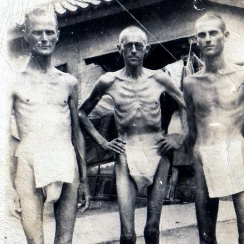 Trois hommes se tiennent devant un bâtiment. Ils sont émaciés et ne portent rien d'autre qu'un pagne.