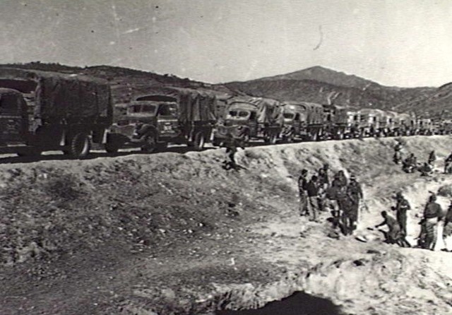 Un groupe de véhicules militaires sur une large route surélevée. Des soldats se tiennent au bord de la route.