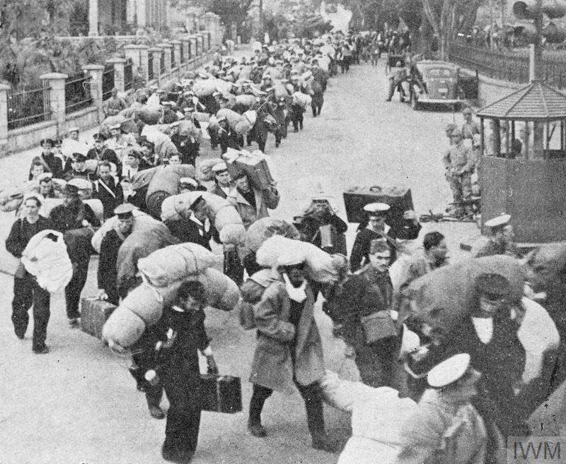 Un groupe important d'hommes en uniforme marche en file indienne. Ils portent de grands sacs et des valises. Un groupe de soldats japonais les observe à proximité.