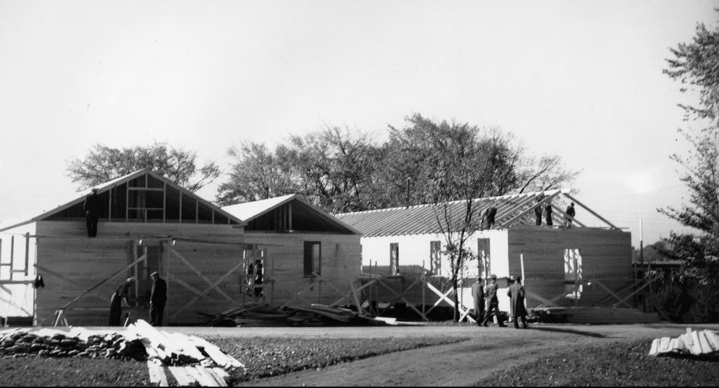 Trois bâtiments sont en cours de construction. Des hommes se tiennent à proximité, tandis que d'autres travaillent sur le toit du bâtiment situé à l'extrême droite.