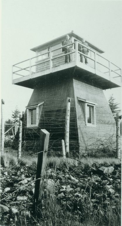 une petite tour en bois. De la barbelée entoure sa base. Deux hommes en uniforme se tiennent sur une plate-forme près du sommet de la tour.